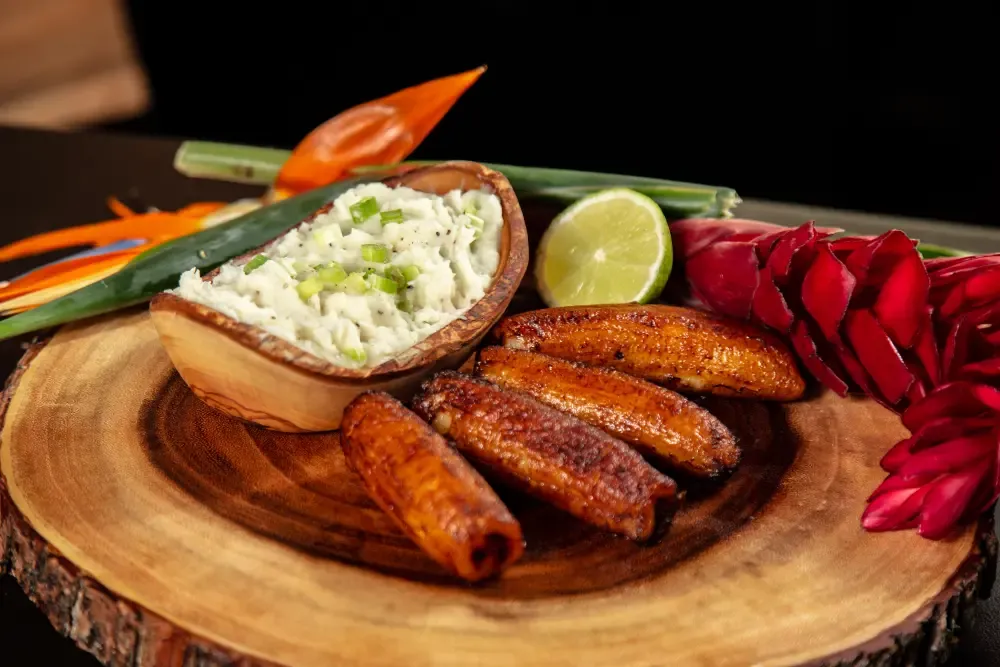 Golden-brown candied bananas and a bowl of mashed white food topped with chives on a rustic wooden board, garnished with a lime wedge and tropical flowers.