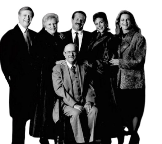 Formal multi-generational family portrait of six smiling individuals, with a man seated and five others standing behind him.