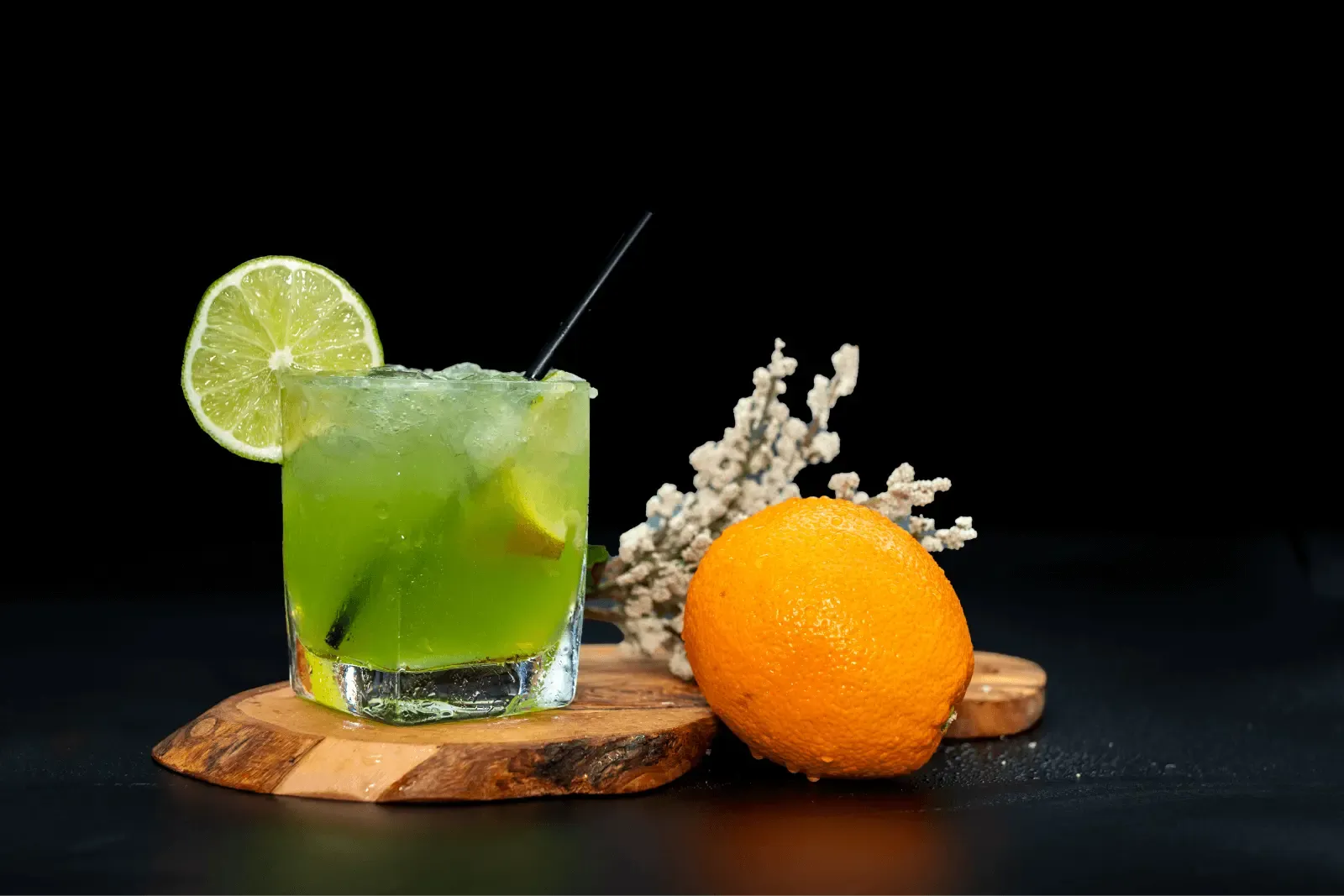 A vibrant green cocktail with a lime garnish and straw on a rustic wooden base, next to a fresh orange and a white decorative branch, against a black background.