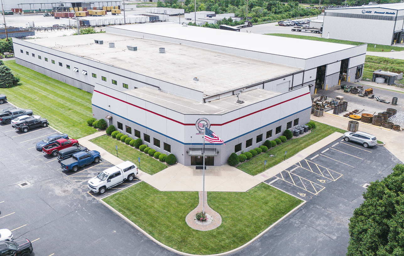 Aerial view of the Arnette Pattern Company factory with an American flag flying at the main entrance, surrounded by a parking lot and green landscaping.