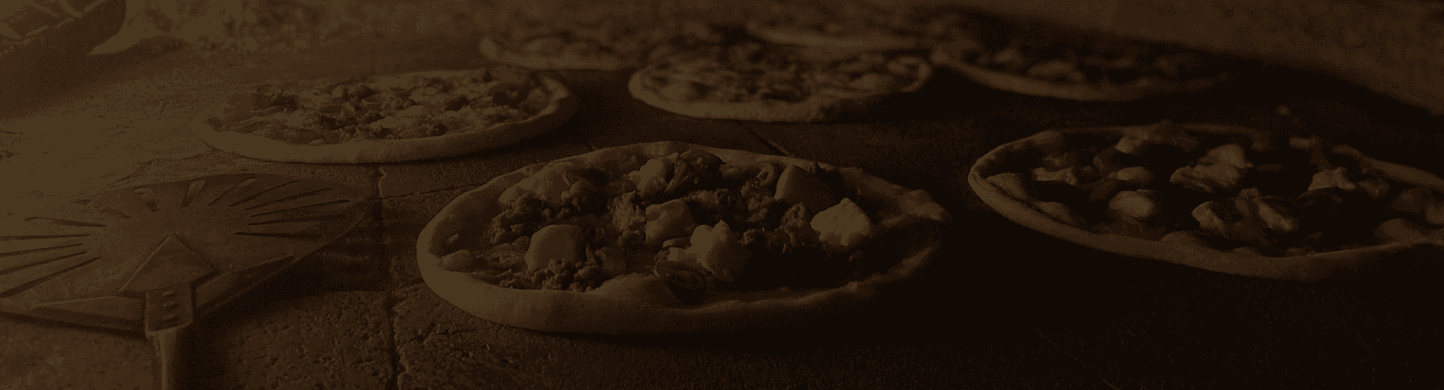 Uncooked pizzas with assorted toppings like cheese, tomato, and meat, arranged on a stone surface next to a pizza peel, in a dim, rustic setting.