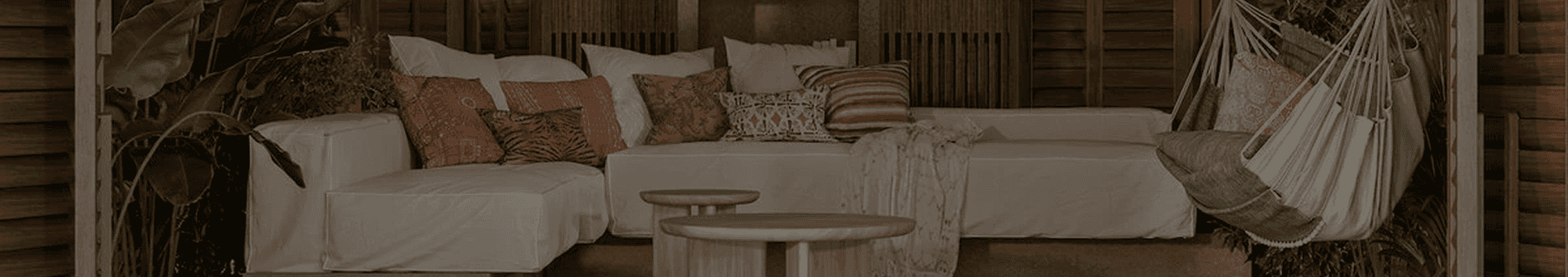 An inviting outdoor lounge with a long white modular sofa, an assortment of patterned pillows, wooden coffee tables, and a hanging chair, set against lush greenery and wooden slat walls.