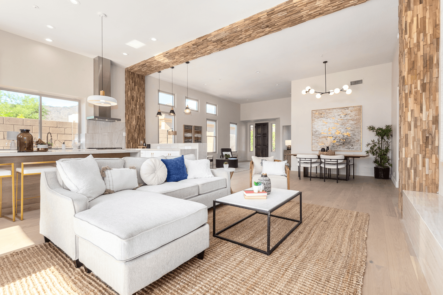 Bright, open-concept living room with a large sectional sofa, modern kitchen island, and dining area, all featuring light wood floors and decorative wood accents.