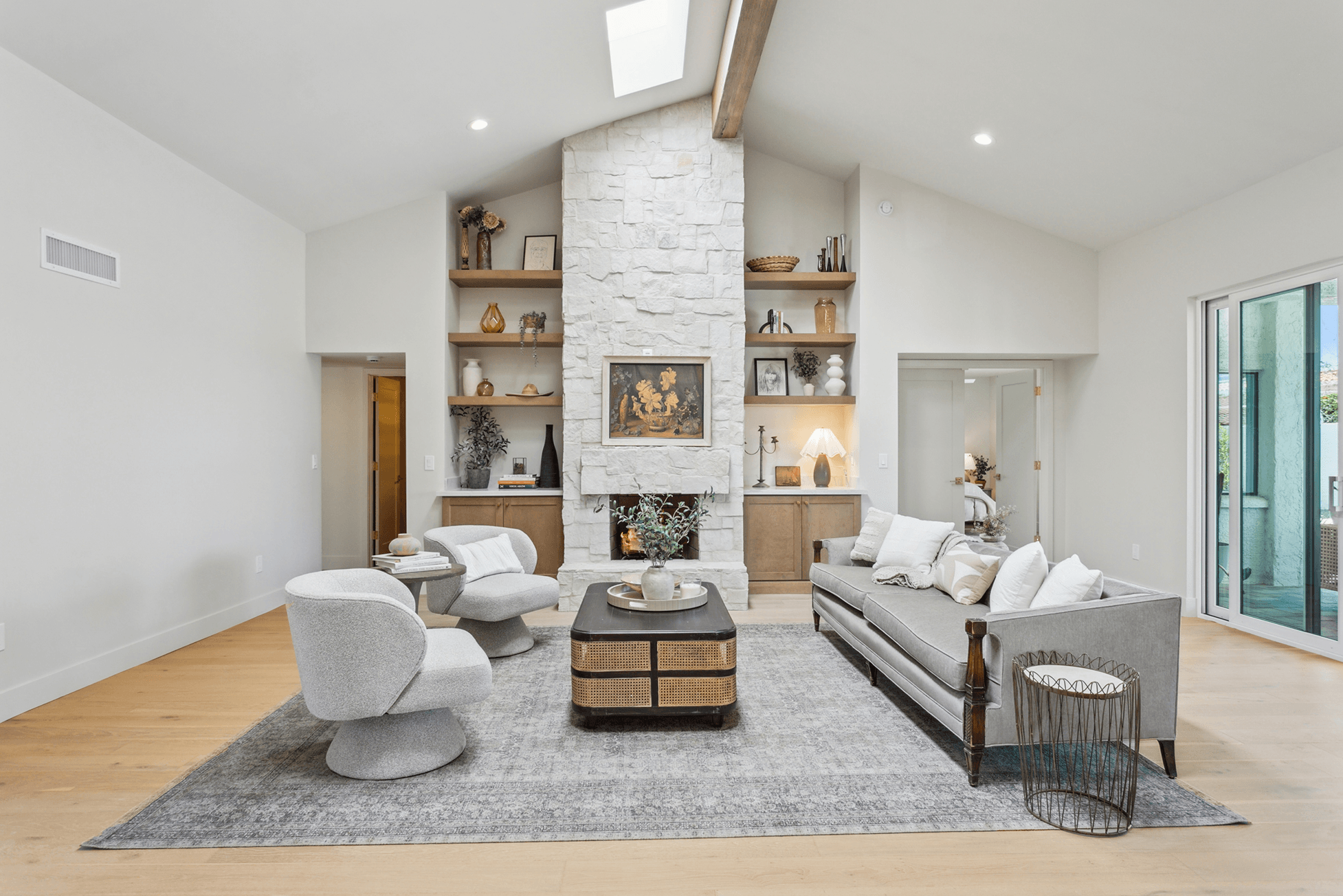 A bright, modern living room with a white stone fireplace, flanked by built-in wood shelves, light wood floors, a gray area rug, and contemporary seating.