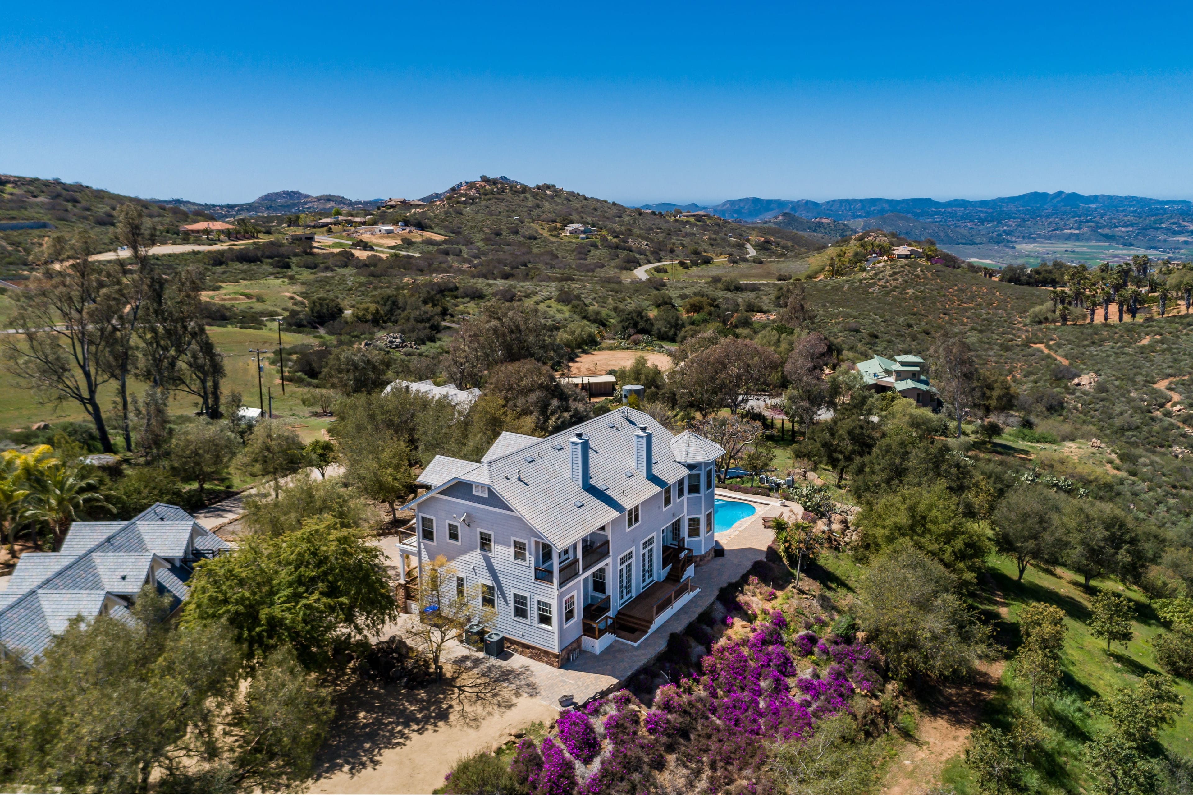 Aerial view of a light-blue house with a swimming pool, nestled among rugged green hills and distant mountains under a clear blue sky.