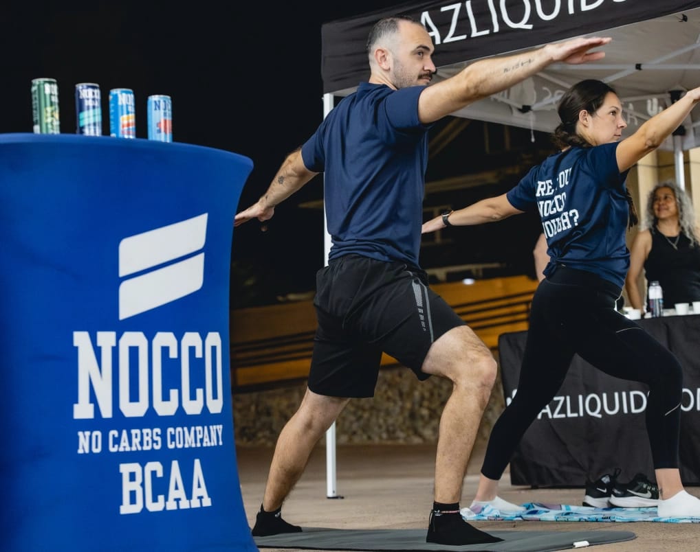 Two people in athletic wear perform a yoga pose outdoors, with a NOCCO branded table featuring energy drinks and a tent in the background.