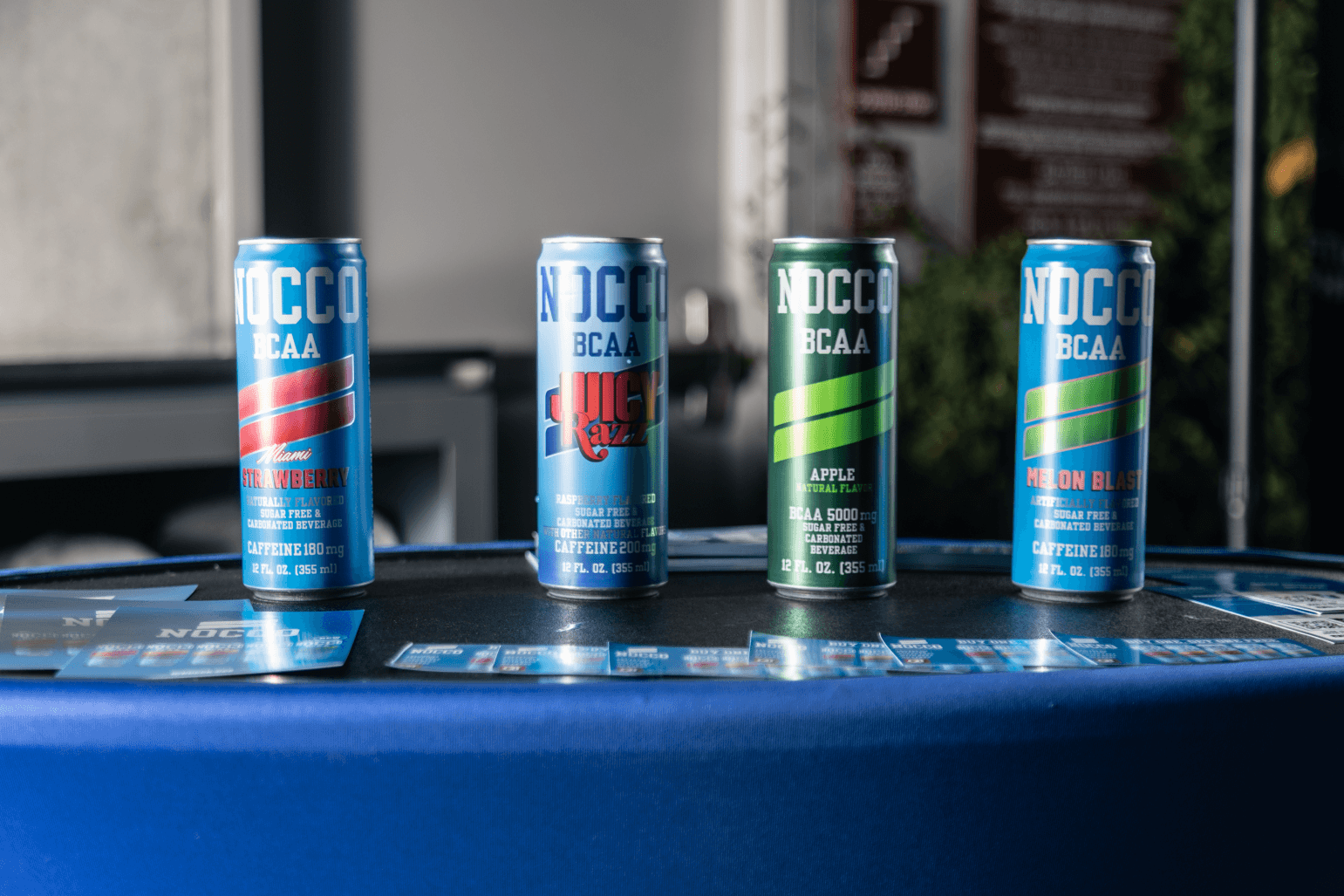 Four NOCCO BCAA sugar-free drinks, including Miami Strawberry, Juicy Razz, Apple, and Melon Blast flavors, displayed with promotional cards.