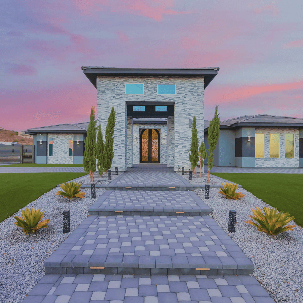 A modern, stone-clad luxury house entrance at dusk, with ornate double doors and a tiered paver pathway. Tall evergreen trees and low plants flank the path, all set against a vibrant pink and purple sky.