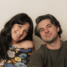 Smiling woman with dark hair and a floral top next to a man with graying hair and a green sweater, both looking at the camera.