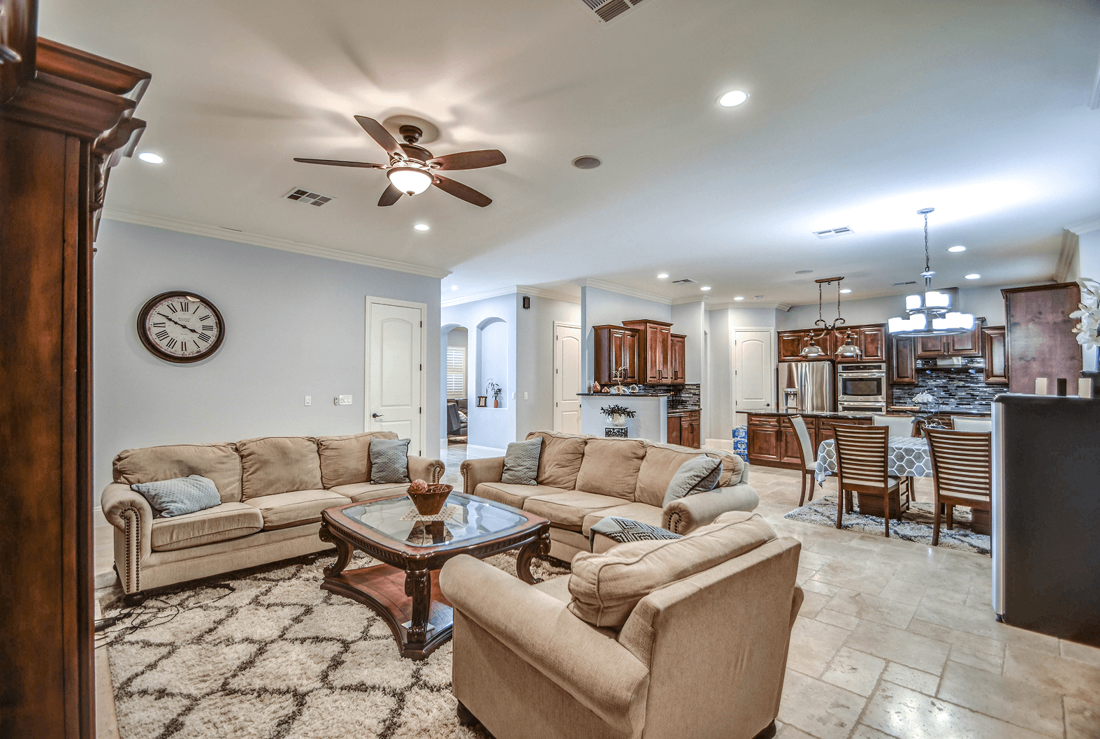 A spacious open-concept living room featuring light-colored sofas, a glass coffee table, and a patterned rug, connecting to a modern kitchen with dark wood cabinetry and a dining table.