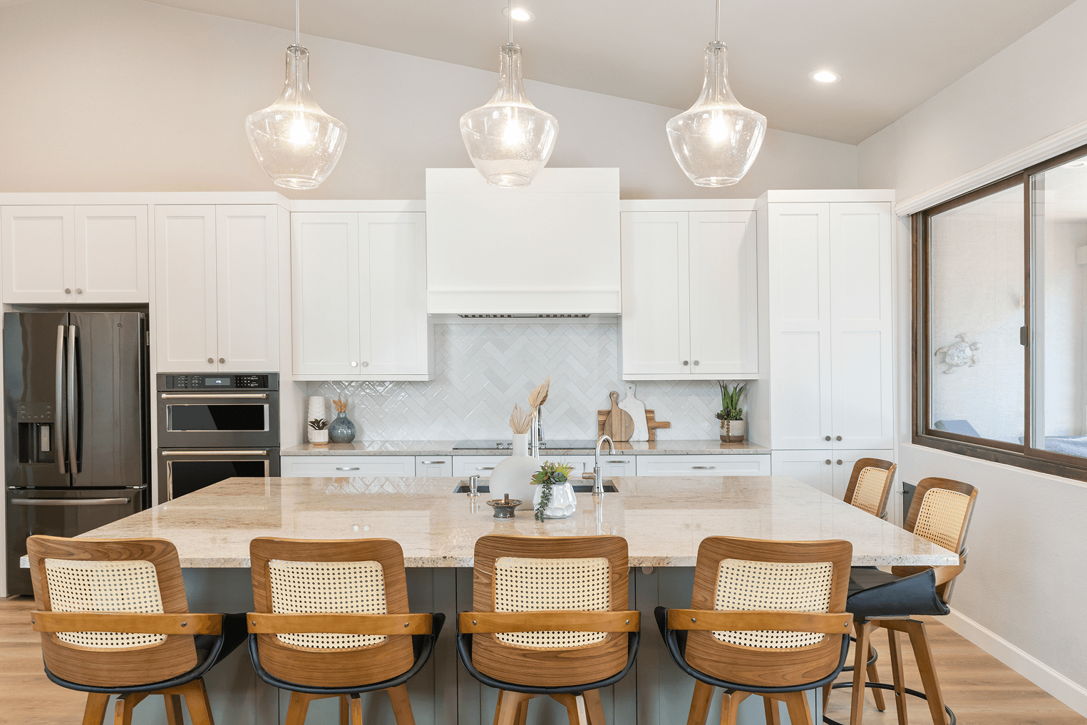 A modern white kitchen with a large granite island, five cane-back bar stools, stainless steel appliances, a herringbone tile backsplash, and three clear pendant lights.