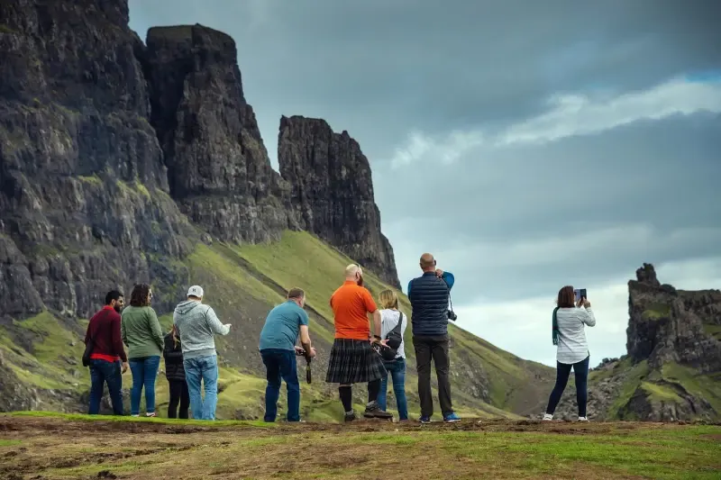 A group of people stand on a grassy hill, admiring a dramatic landscape of towering, jagged cliffs and green slopes under a cloudy sky. One person wears a kilt.
